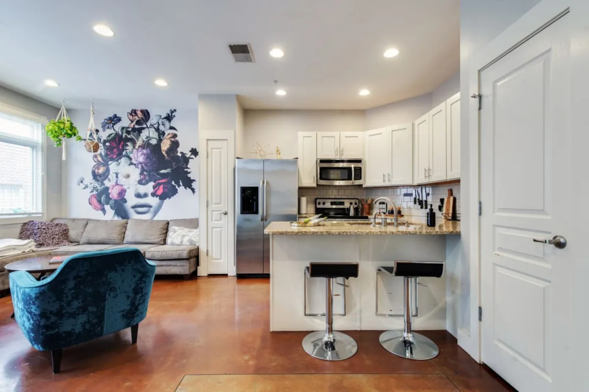 A stylish open-concept space featuring a large floral mural as the artistic centerpiece, with a kitchen island in the foreground and a comfortable sofa nearby. The space is bathed in natural light, ideal for social gatherings and culinary activities.