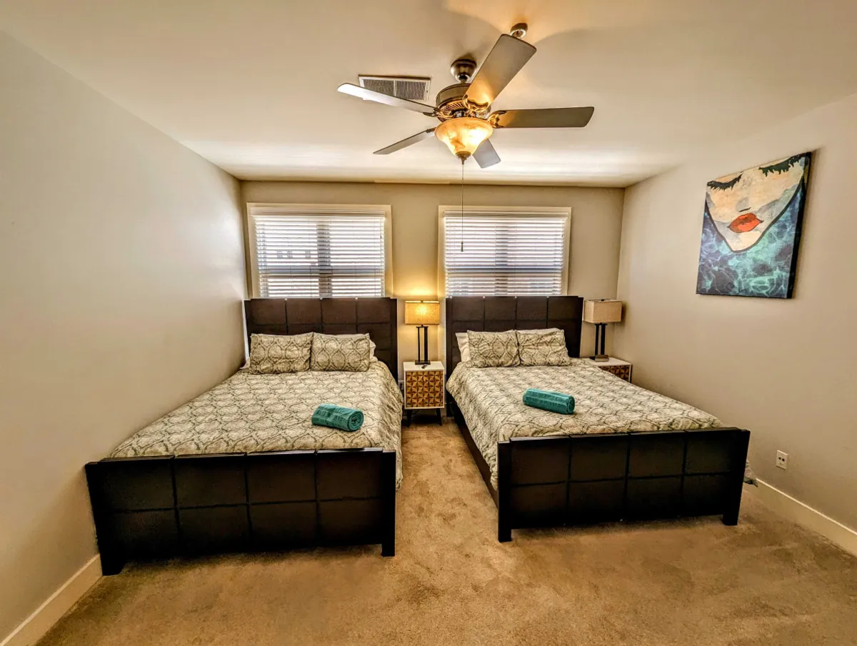 A welcoming bedroom featuring two queen beds with decorative bedding, a ceiling fan overhead, and tasteful artwork on the wall. The space is designed for both restful sleep and casual lounging, perfect for families or friends sharing a room.