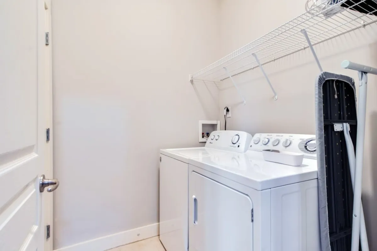 A tidy laundry area featuring a modern washing machine, dryer, ironing board, and organized shelves for laundry essentials. The space is well-lit and designed for practical, everyday use.