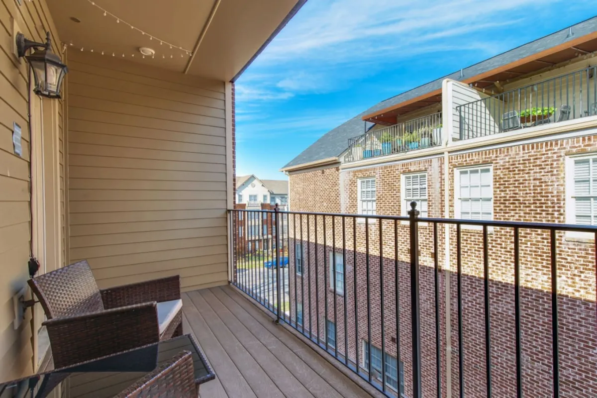 A private balcony with a sturdy railing, comfortable outdoor seating, and neighboring buildings in view. The scene features someone relaxing with a book and another enjoying a morning coffee, set against a calm, inviting atmosphere. The balcony is an intimate space, elevated above the city bustle, perfect for quiet moments or casual entertaining.