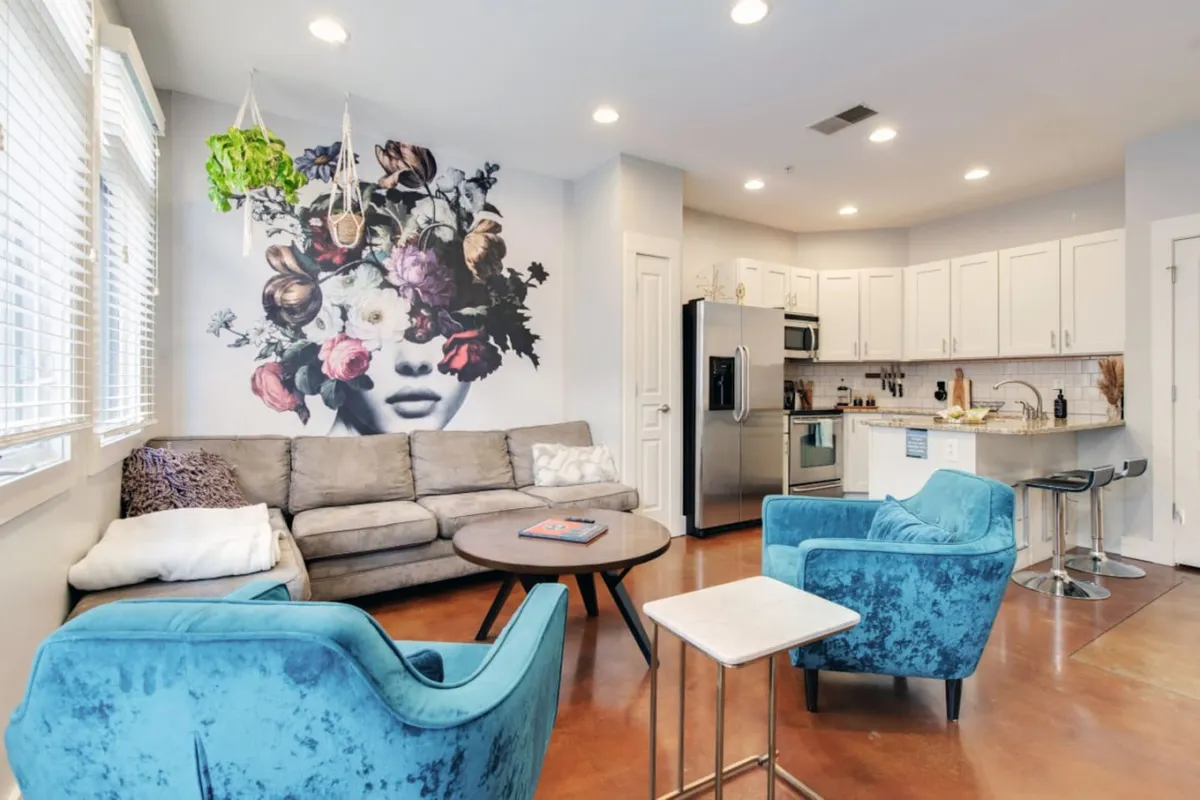 A spacious living area with a striking large wall mural featuring a face adorned with flowers, paired with blue accent chairs, a modern kitchen in the background, and a coffee table with a book. The overall vibe is vibrant and welcoming, ideal for gathering and relaxing after neighborhood adventures.