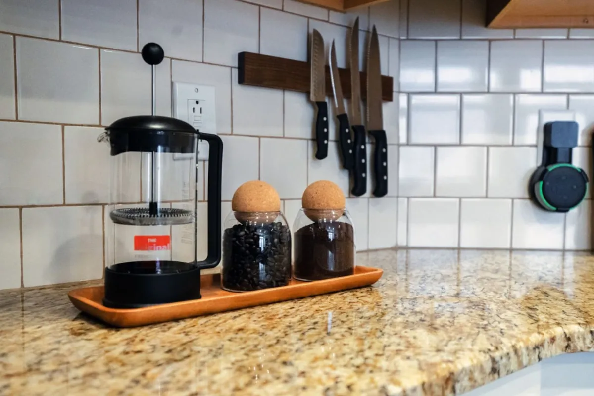 A cozy kitchen corner showcasing a French press coffee maker, glass jars with cork lids, and a set of kitchen knives. The setup suggests warmth and comfort, making it easy for guests to brew fresh coffee and prepare meals during extended stays.