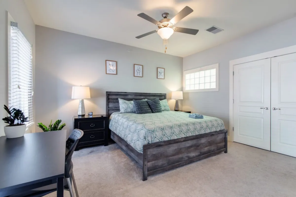 A serene bedroom with a neatly made bed, a nightstand, a small desk, and a window letting in soft natural light. The room feels calming and private, offering space for both rest and quiet activities.