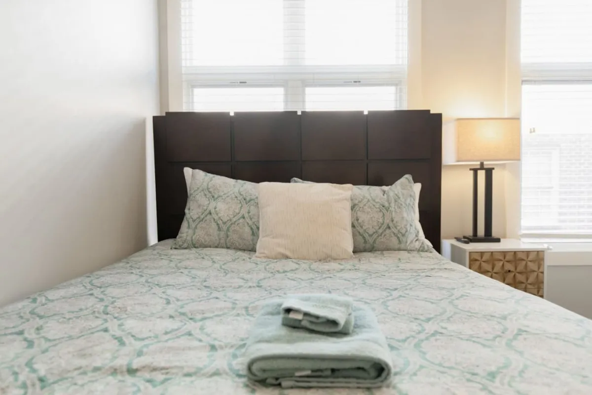 This image showcases a bed with patterned bedding, decorative pillows, and a lamp on the nightstand, set near a window. The tranquil atmosphere and presence of the window evoke the experience of enjoying a relaxing morning or unwinding after exploring the neighborhood, especially with a cup of coffee in hand.