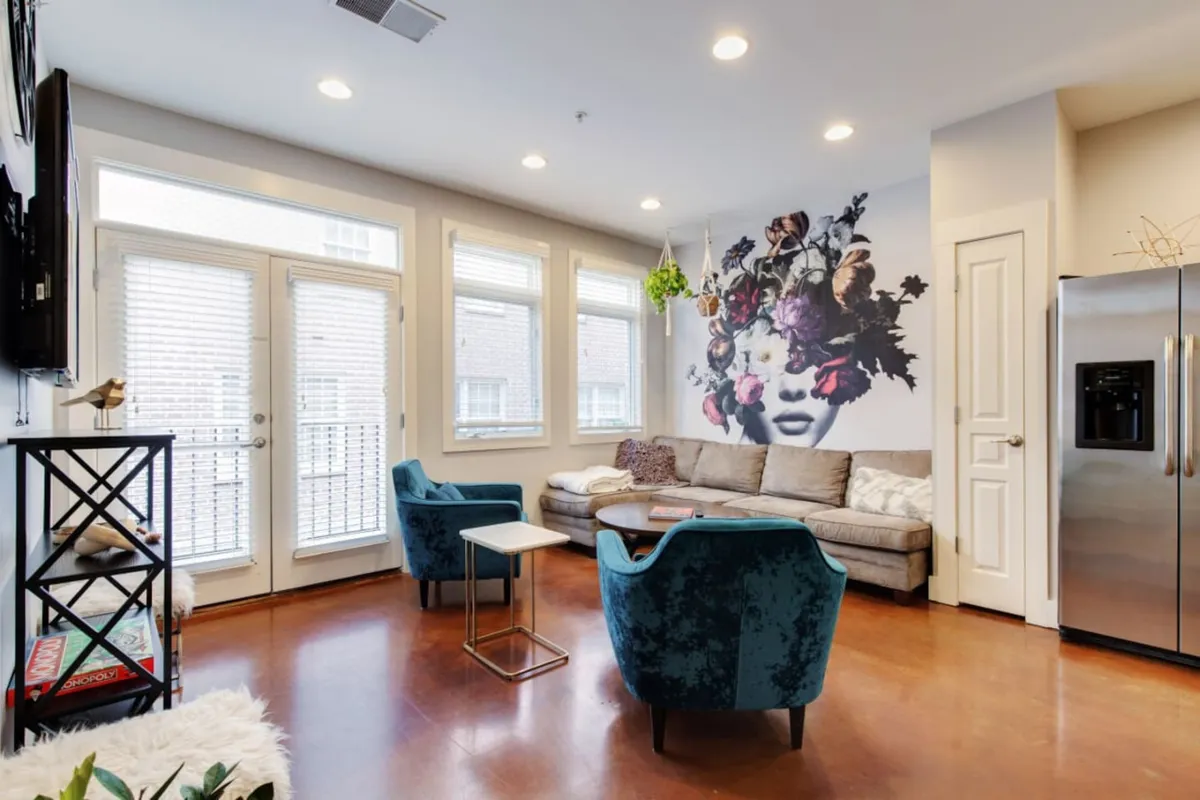 A stylish living area featuring a striking mural of a woman's face adorned with flowers, a modern couch, colorful armchairs, and a glass table. Bright natural light fills the room, highlighting an inviting, creative atmosphere ideal for relaxing or informal meetings.