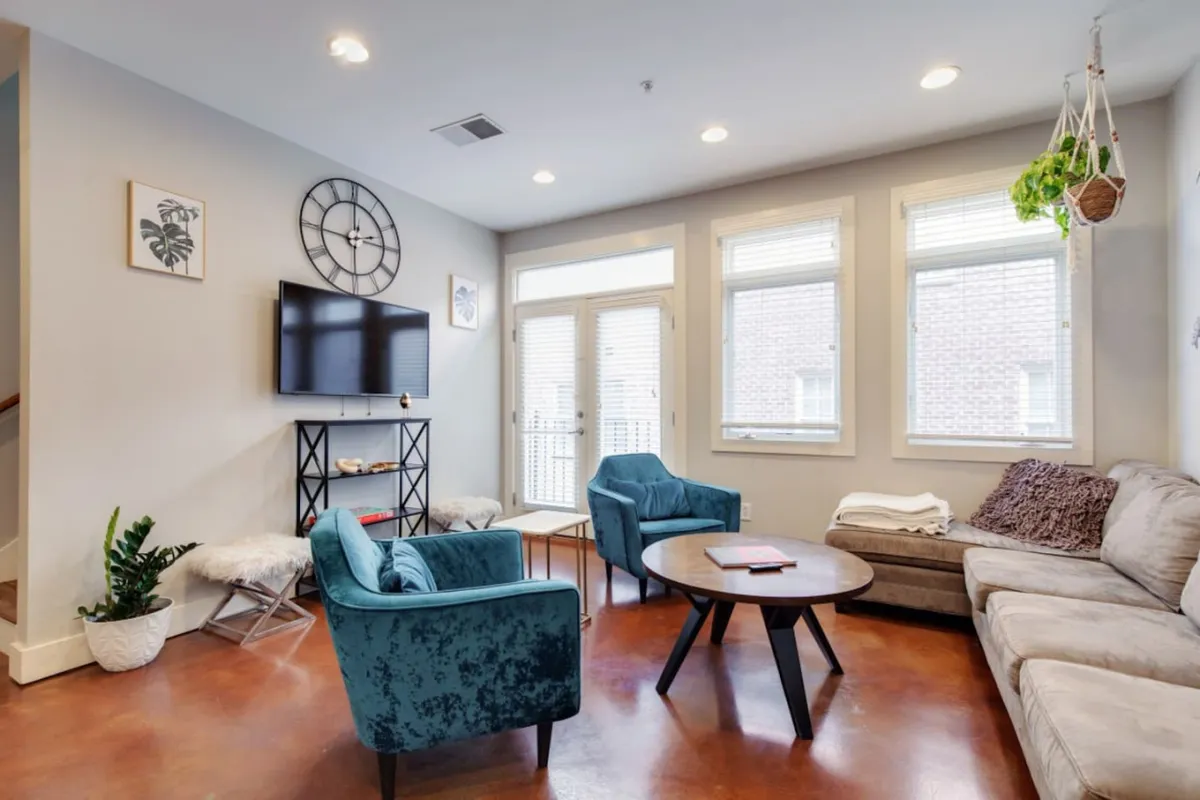 A cozy den with a TV unit, comfy blue chairs, a coffee table, and a large window letting in natural light. The window view suggests a connection to the quiet, historic neighborhood outside while the room itself is set up for relaxation or family time.